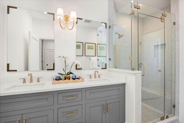 Modern bathroom with a double vanity, gold fixtures, large mirrors, and a glass-enclosed shower; decor includes a vase and framed art.