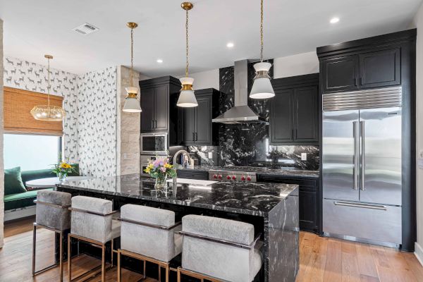 A modern kitchen with black cabinets, marble countertops, pendant lights, a stainless steel fridge, and a seating area with gray chairs.