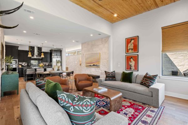 A modern living room with colorful sofas, art pieces, a wooden ceiling, and an open kitchen and dining area in the background.