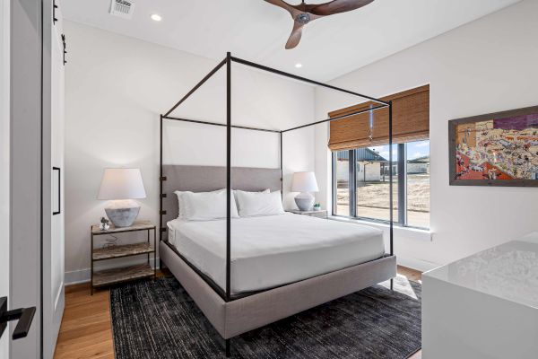 A modern bedroom with a canopy bed, two lamps, a dark rug, window with blinds, and colorful wall art is shown.