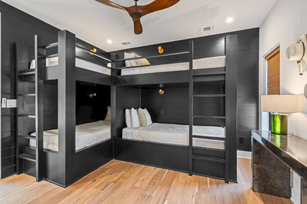The image shows a modern room with black wooden quad bunk beds, a ceiling fan, and a side table with a lamp.