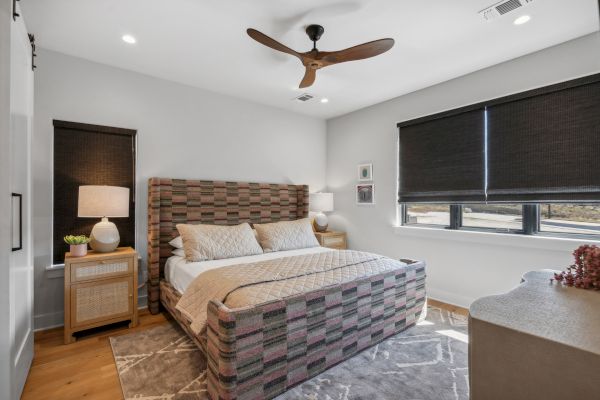 A modern bedroom features a patterned bed, bedside tables with lamps, a ceiling fan, a rug, and blackout blinds on the windows.