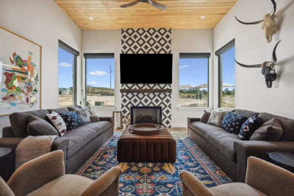 A modern living room with two sofas, patterned rug, decorative fireplace, wall art, and animal head decor. Large windows offer natural views.