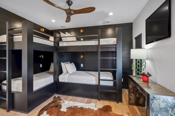 A modern bedroom with two bunk beds, a cowhide rug, ceiling fan, TV, and a lamp on a dresser. Walls have dark wood paneling.