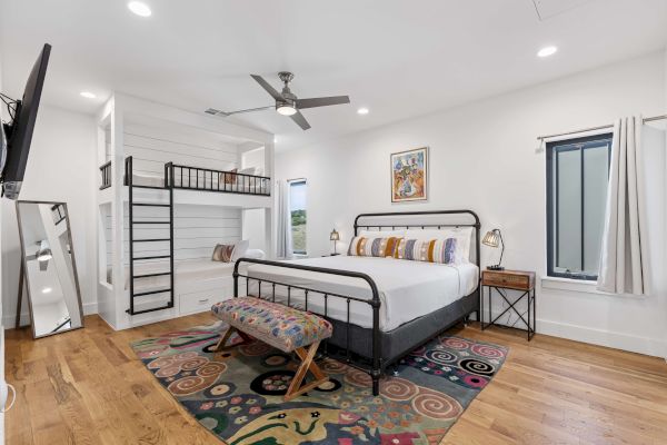 A bedroom with a double bed, bunk bed, decorative rug, bench, wall art, and modern decor, featuring wood floors and a ceiling fan.