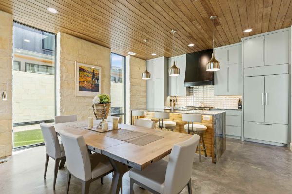A modern kitchen and dining area features wooden ceiling, light fixtures, and neutral tones, with a dining table and kitchen island.