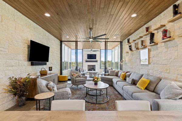 A modern living room with a large gray sectional sofa, stone walls, TV, and ceiling fan, featuring decorative shelves and large windows.