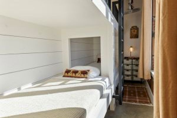 The image shows a cozy bunk bed setup in a narrow room with white walls, pillows, a patterned rug, and curtains.