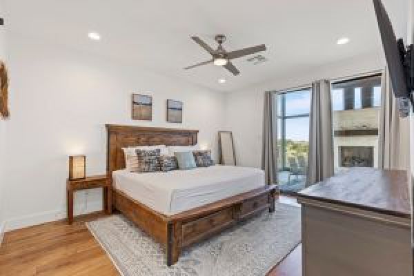 A bedroom with a wooden bed, pillows, a ceiling fan, a dresser, a rug, lamps, and large windows with curtains.
