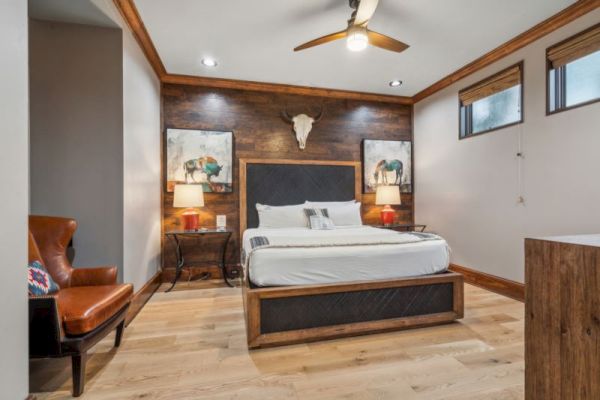 The image shows a bedroom with a wooden floor, a bed with two paintings above, two side tables with lamps, and a chair in the corner.