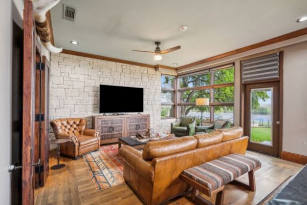 A cozy living room with leather sofas, a large TV, a stone accent wall, and large windows overlooking greenery and water outside.