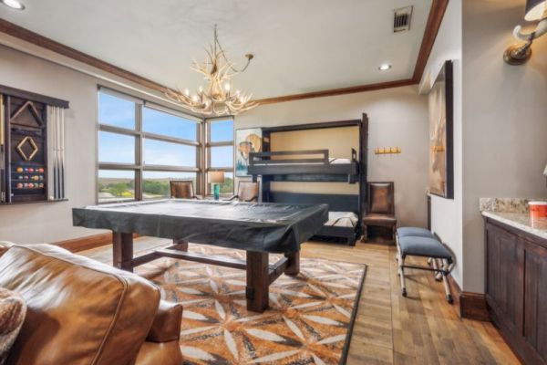 A cozy room with a pool table, bunk beds, rustic chandelier, and large windows offering a scenic view.