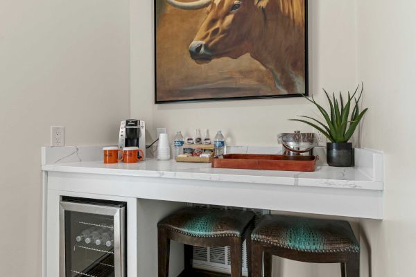 A small beverage station with stools, a mini-fridge, coffee machine, and wall art featuring a large animal in a cozy corner setup.