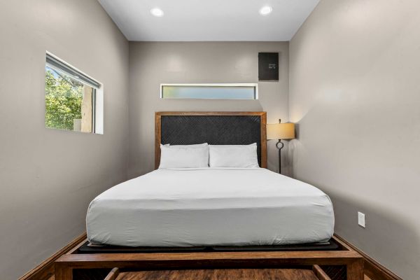 A neatly made bed with white linens is centered in a small room, featuring a wooden headboard and two windows on the walls.