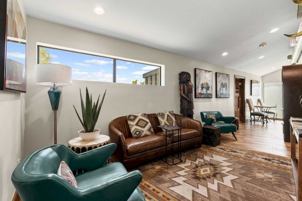 A stylish living area with leather sofa, teal chairs, decorative plants, art, and a large window, exuding a cozy, modern vibe.