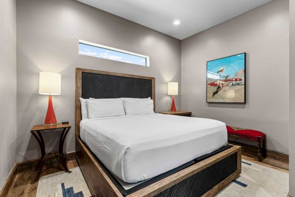 A modern bedroom with a bed, side tables, lamps, and artwork featuring an airplane. Walls are light gray and the room is well-lit.