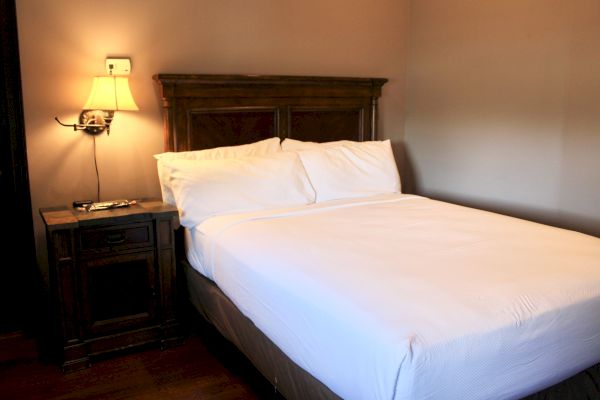 A neatly made bed with white sheets is beside a wooden nightstand with a lamp on, creating a cozy bedroom atmosphere.