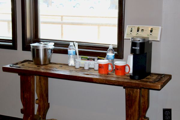A wooden table with a coffee maker, mugs, water bottles, and condiments set up against a window near an electrical outlet.