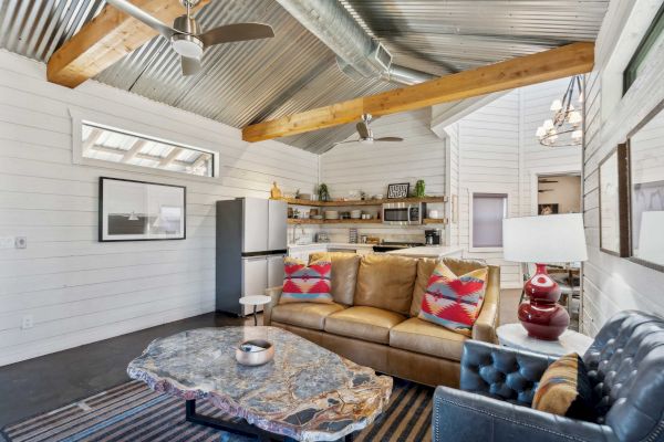 A cozy living room with a sofa, colorful cushions, modern furnishings, and a kitchen area under a high ceiling with exposed beams.