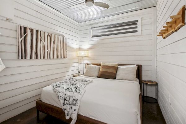A cozy bedroom features a bed with pillows and a blanket, a wall art piece, a lamp, and a corrugated metal ceiling for a rustic touch.