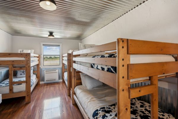 The image shows a room with four wooden bunk beds, cow print bedding, wooden floors, and a window offering a view outside.
