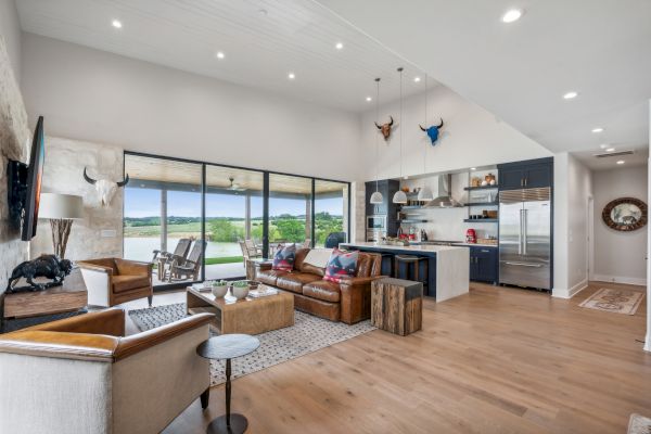 The image shows a modern open-plan living area with a kitchen, wooden floors, large windows, and stylish furniture in neutral tones.