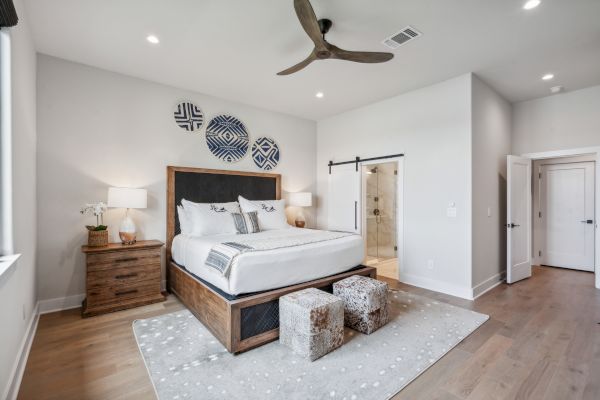 A modern bedroom with a wooden bed, decorative wall art, and a ceiling fan. The room has bedside tables and plush stools at bed's foot.