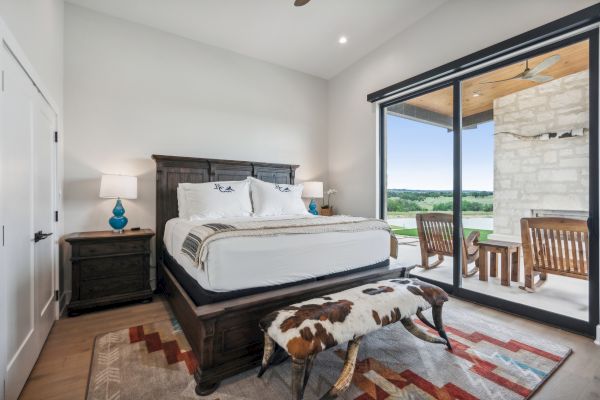 A cozy bedroom featuring a large bed, cowhide bench, and a view of a patio with chairs and greenery outside a sliding glass door.