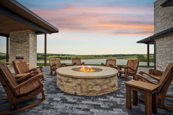 An outdoor patio with a stone fire pit surrounded by wooden chairs, flanked by stone buildings, and overlooking a scenic landscape at sunset.