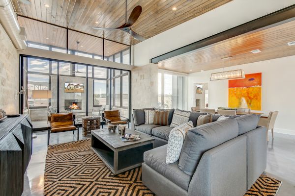 A modern living room with a grey sectional, geometric rug, and large windows. There's a fireplace, ceiling fan, and vibrant artwork.
