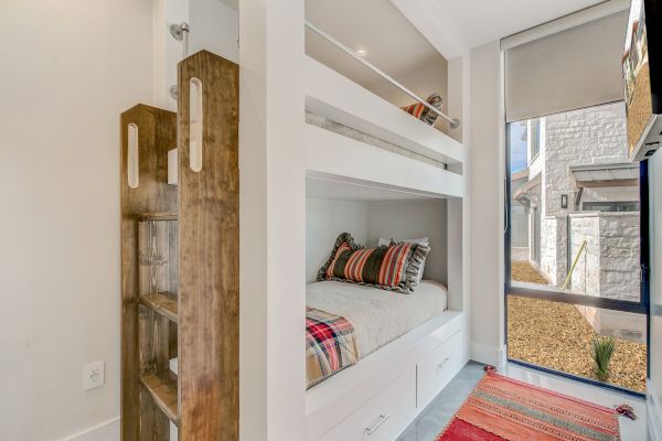 The image shows a small bedroom with wooden bunk beds, colorful pillows, a window with a shade, and a red rug on the floor.
