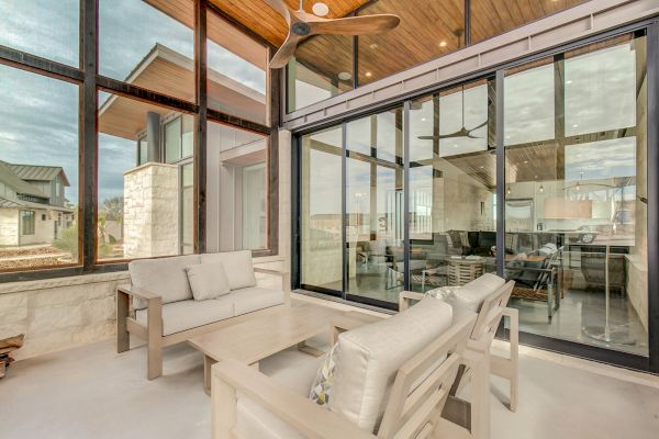 A modern indoor-outdoor space with glass walls, wooden ceiling, light furniture, and large windows with a ceiling fan above.