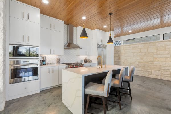 Modern kitchen with white cabinets, stainless steel appliances, pendant lights, an island with chairs, and a wooden ceiling.