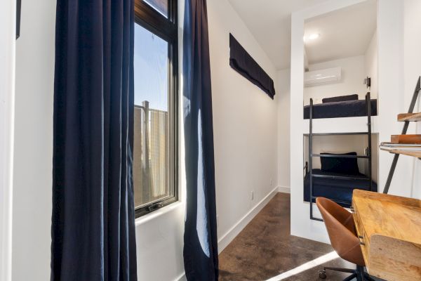 A small, modern room with a bunk bed, desk, chair, and windows with black curtains. There's an air conditioner on the wall.