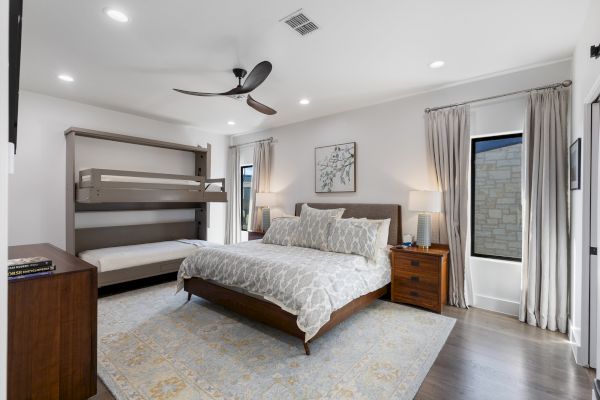 A modern bedroom with a bed, a fold-out bunk, nightstands, lamps, a ceiling fan, and curtains. Neutral colors dominate the decor.