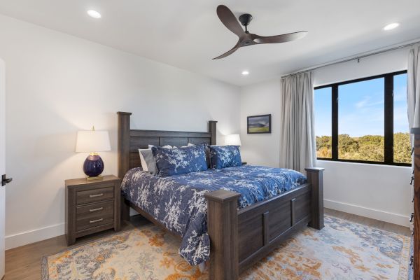 A bedroom with a wooden bed, blue bedding, two nightstands, a ceiling fan, a large window, and a patterned rug.