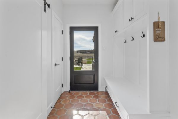 The image shows a hallway with a tiled floor, a black door, white walls, coat hooks, and seating bench with storage underneath.