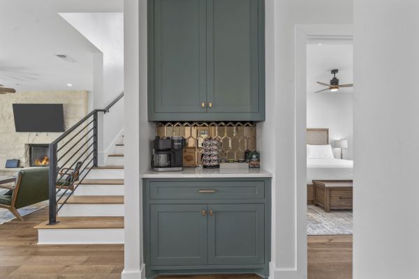 A cozy kitchen nook features a coffee station with cabinets, next to a staircase and living room; a bedroom is visible through a doorway.