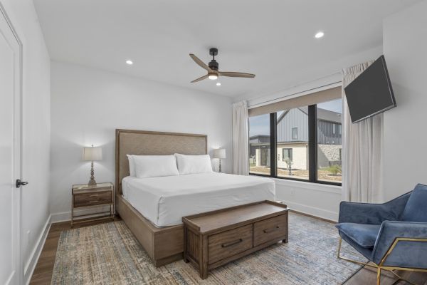 A modern bedroom with a bed, nightstands, ceiling fan, large window, TV, and a blue chair on a patterned rug.