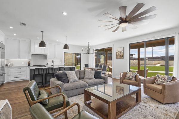 A modern living room with a kitchen, sofas, armchairs, a ceiling fan, and large windows overlooking a patio and yard.