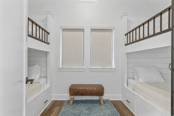 The image shows a small room with two sets of bunk beds, rolled-up blinds, a brown bench, and a blue rug on a wooden floor.