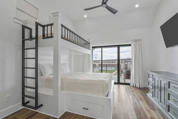 A modern bedroom features a bunk bed, ceiling fan, wall-mounted TV, dresser, and large glass doors opening to a view outside.