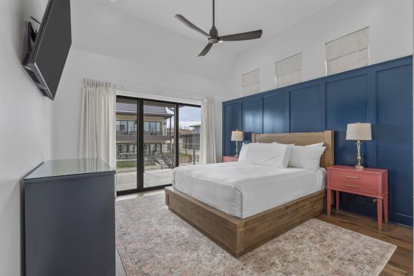 A modern bedroom with a wooden bed, blue accent wall, large windows, pink side tables, ceiling fan, and wall-mounted TV.