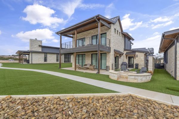 A modern house with stone and wood accents, large windows, a covered balcony, and a patio surrounded by artificial grass and pathways.