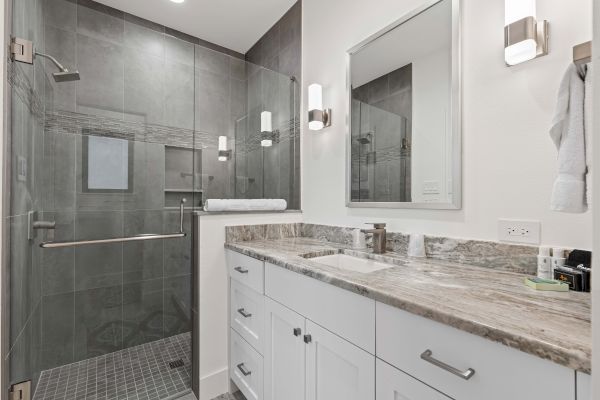 A modern bathroom with a glass-enclosed shower, marble countertop, and white cabinets, illuminated by wall lights above the vanity mirror.
