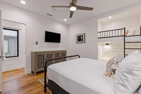 A modern bedroom with a bed, bunk beds, TV, ceiling fan, and wooden flooring. Wall decor and soft lighting add a cozy feel.