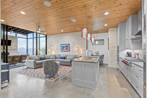 The image shows a modern open-concept living room and kitchen with a wooden ceiling, large windows, grey cabinets, and stylish decor.