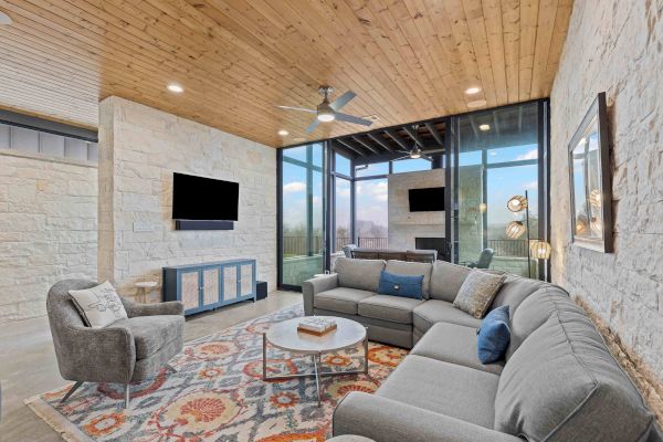 A modern living room featuring a sectional sofa, armchair, TV, and a large rug. It has glass walls, wooden ceiling, and outdoor view.