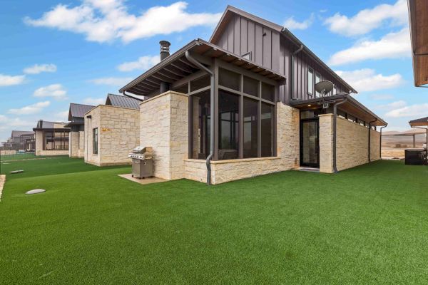 A modern house with stone walls and large windows, surrounded by artificial grass, under a bright blue sky with white clouds.